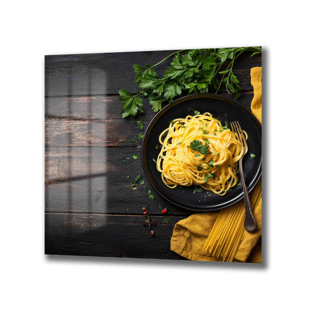 Koyu kahverengi ahşap bir masa üzerinde, siyah tabakta servis edilmiş, taze maydanozla süslenmiş iştah açıcı sarı spagetti tabağı. Yanında bir çatal, sarı bir kumaş peçete ve kuru spagetti demetleri bulunuyor. Masa üzerinde ayrıca baharat taneleri de serpiştirilmiş.