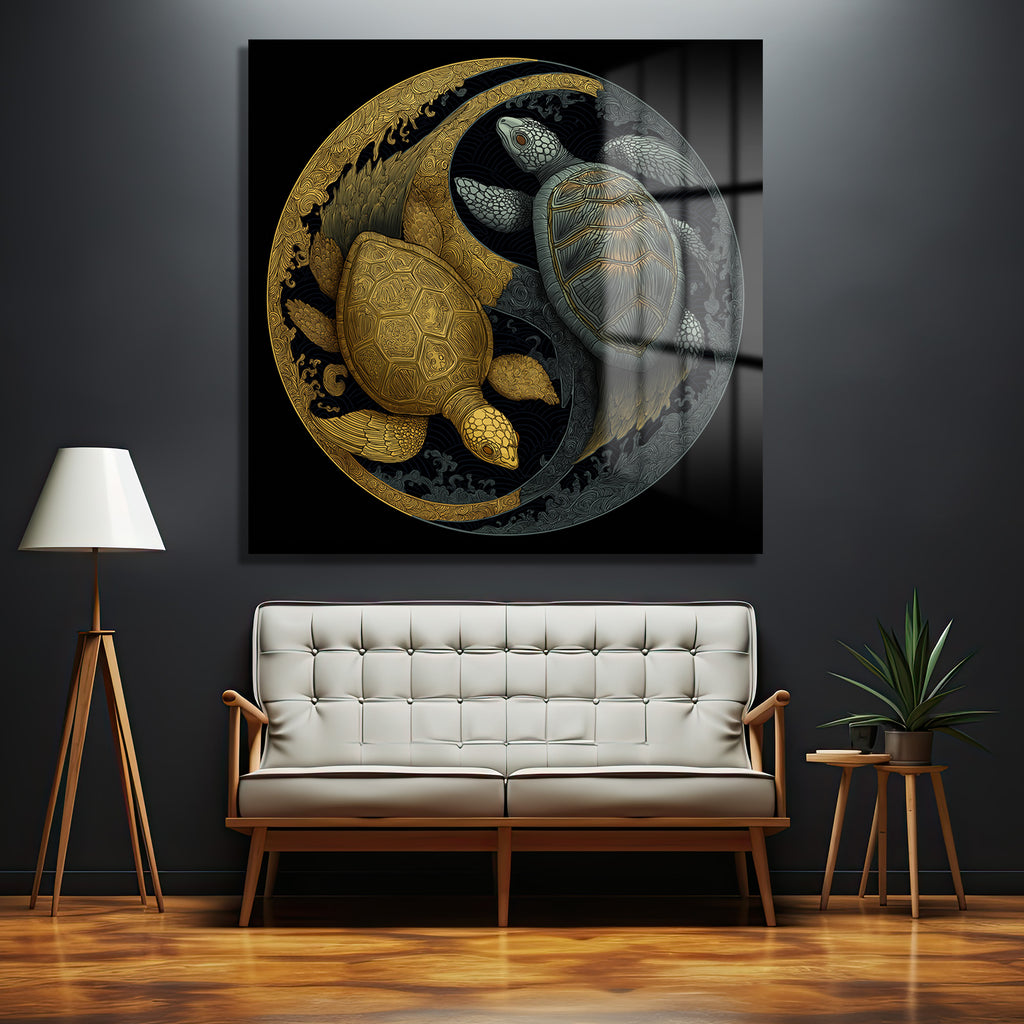 Koyu gri bir duvarda asılı, altın ve gümüş renklerinde iki kaplumbağanın Yin Yang formunda betimlendiği, detaylı desenlere sahip modern bir cam tablo. Kaplumbağalar, siyah bir zeminde, kıvrımlı dalga benzeri motiflerle çevrili. Mekanda beyaz bir kanepe, ahşap bir lambader ve yeşil bir bitki bulunmaktadır.