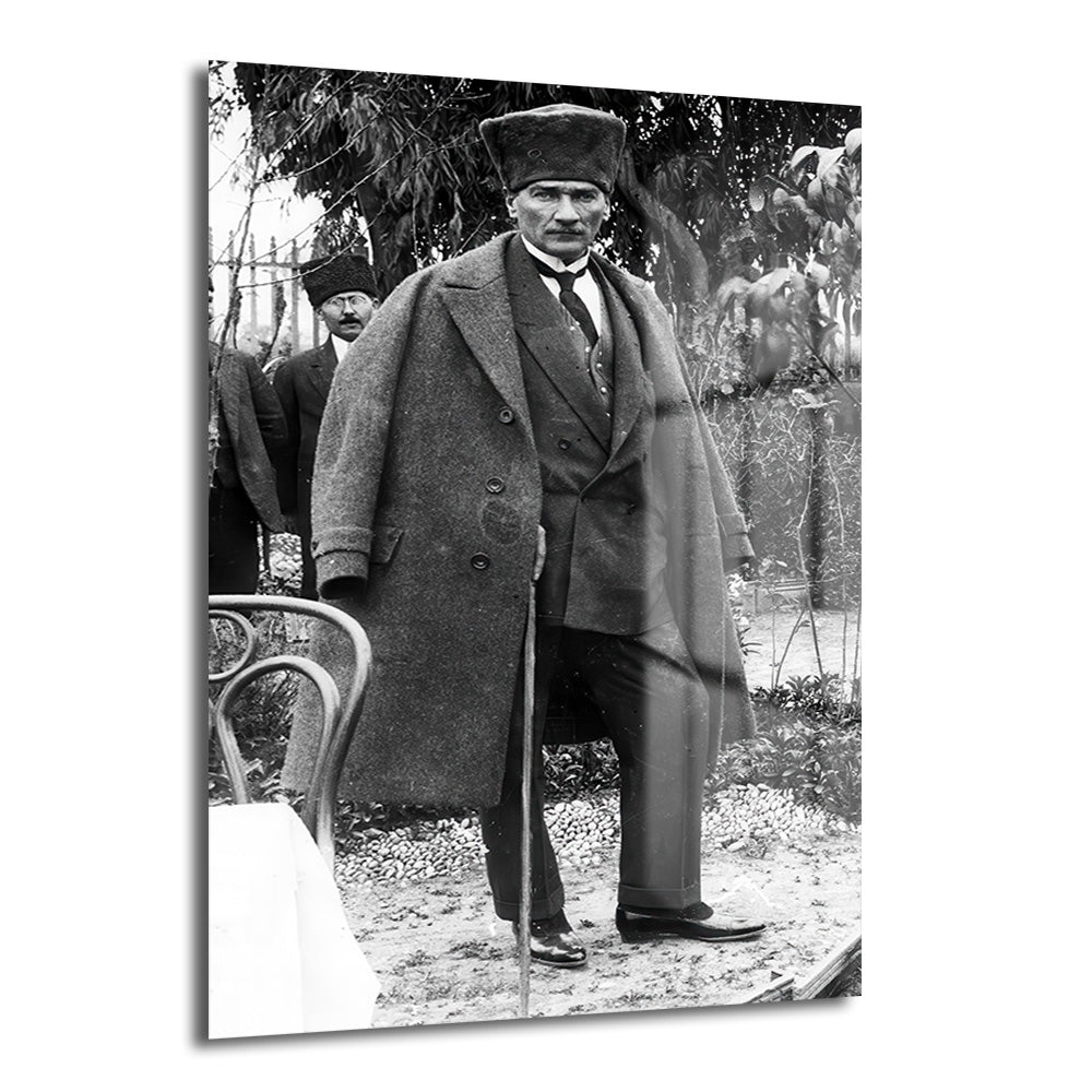 Mustafa Kemal Atatürk'ün şapkalı, pardösülü ve bastonlu olarak bahçede çekilmiş, arkasında bir başka kişinin de göründüğü siyah beyaz, tarihi bir fotoğrafı.