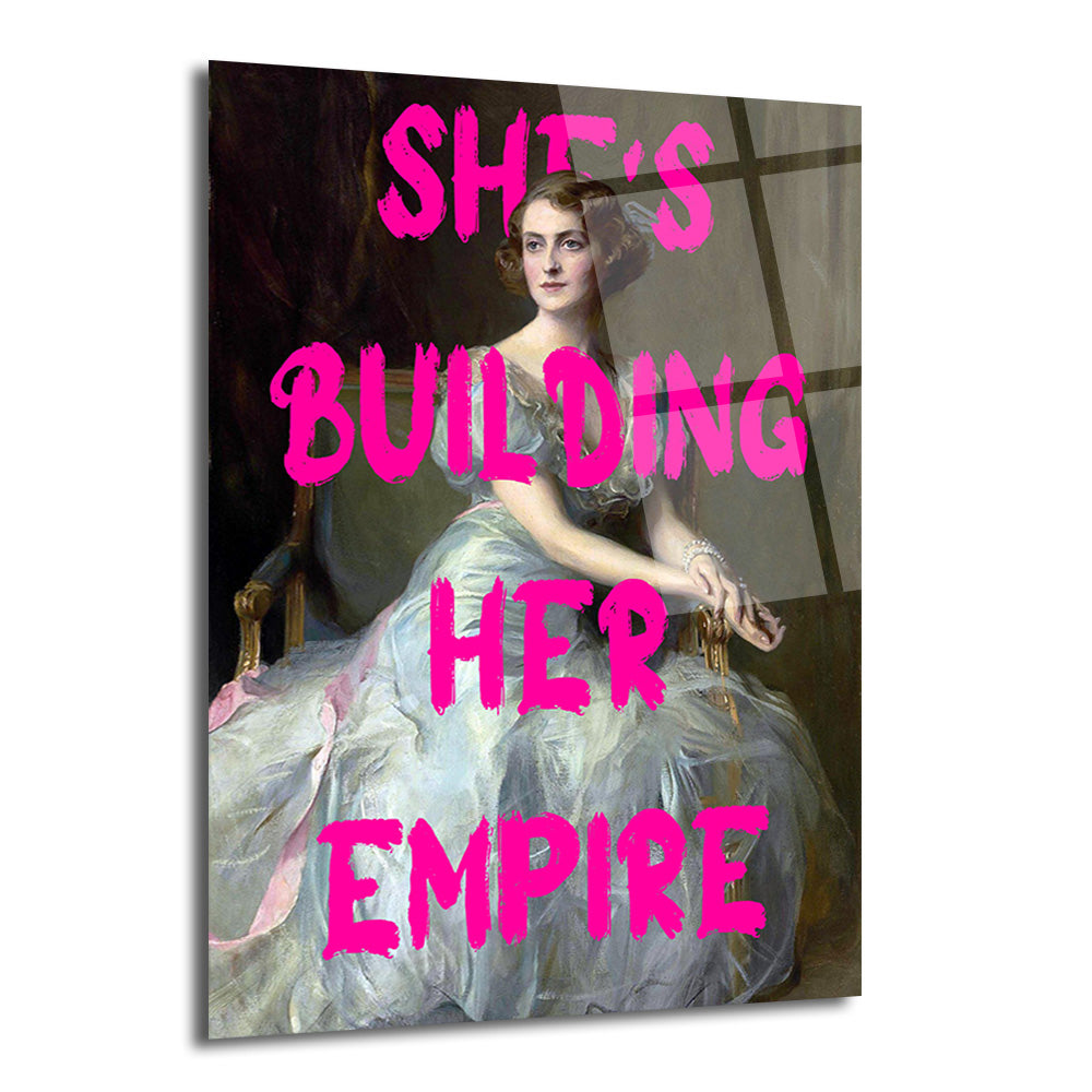 Klasik bir portre üzerine parlak pembe fırça darbesiyle 'SHE'S BUILDING HER EMPIRE' yazısı işlenmiş, şık bir kadının oturduğu cam tablo.